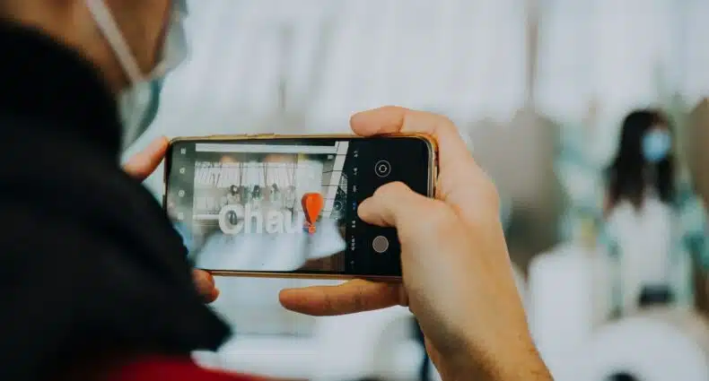 Pessoa branca tirando foto de letras formando a palavra "Chau!" com um ponto de exclamação vermelho. Ao fundo há uma mulher usando máscara. - Foto: Greta Schölderle Møller via Unsplash