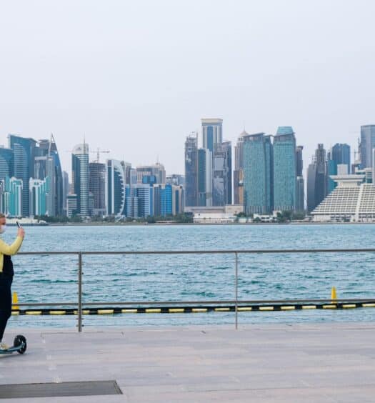 Mulher loira usando macacão jeans e casaco amarelo no canto esquerdo ao lado de um patinete tirando foto da paisagem a sua frente para ilustrar o post sobre chip de celular para Doha. Ao fundo há diversos prédios com o mar em frente. - Foto: Lukhmanul Hakeem via Unsplash