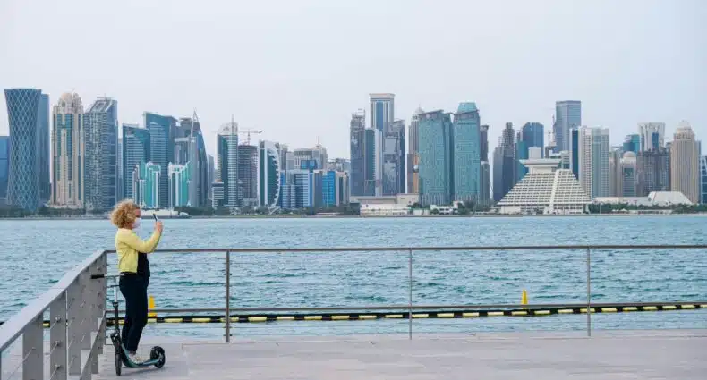 Mulher loira usando macacão jeans e casaco amarelo no canto esquerdo ao lado de um patinete tirando foto da paisagem a sua frente para ilustrar o post sobre chip de celular para Doha. Ao fundo há diversos prédios com o mar em frente. - Foto: Lukhmanul Hakeem via Unsplash
