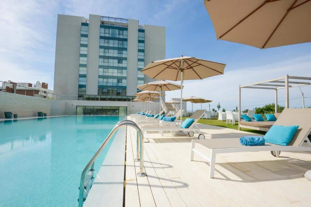 Vista da frente do The Grand Hotel  com piscina e cadeiras com guarda sóis ao lado da piscina. Representa hotéis em Punta Del Este.