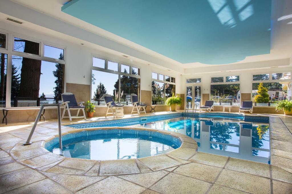 Piscina coberta do Huinid Pioneros Hotel com duas piscinas - uma maior e uma pequena - com cadeiras perto da piscina grande e várias janelas de vidro. Representa hotéis em Bariloche
