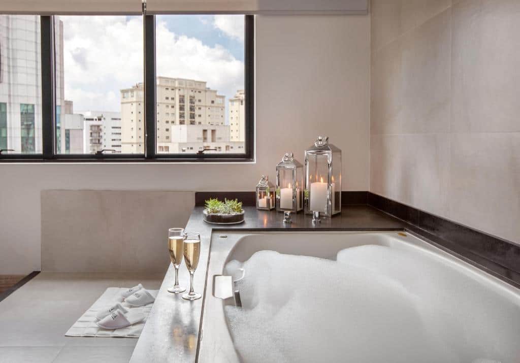 Banheiro do Meliá Ibirapuera com uma banheira de hidromassagem, uma janela ampla com vista para a cidade e alguns itens de decoração