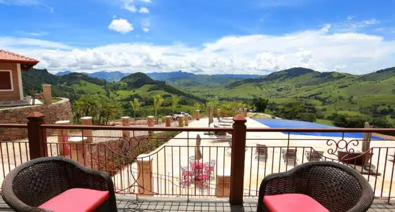 Área externa da Pousada Aldeia Manacás, com uma vista panorâmica de montanhas verdes e céu azul com nuvens brancas. Há também uma piscina com várias espreguiçadeiras em volta e um deck de madeira com duas poltronas em cima, para representar pousadas na Serra da Mantiqueira