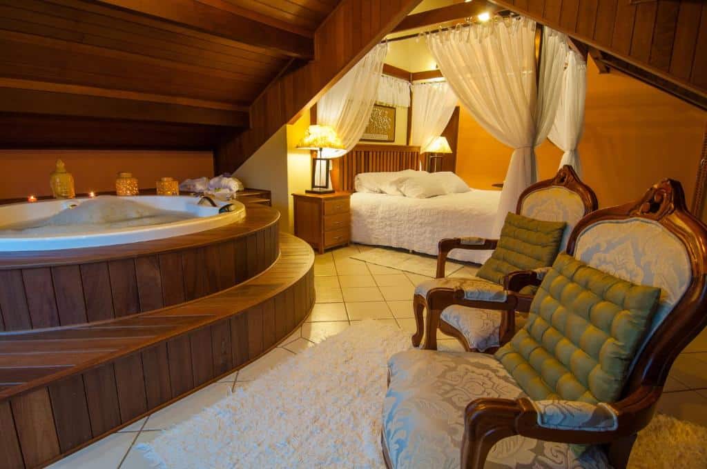 Interior do quarto de uma das pousadas na Serra da Mantiqueira, com teto de madeira, hidromassagem, uma cama de casal e duas poltronas de época com almofadas verdes em cima