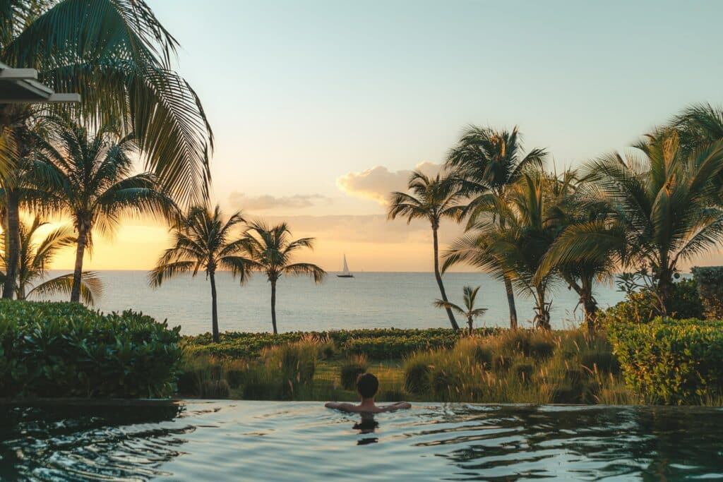 Pessoa com os braços debruçados na borda de uma piscina, olhando para o pôr do sol em uma praia, que tem um barco no horizonte e uma parte costeira com gramado verde e algumas árvores
