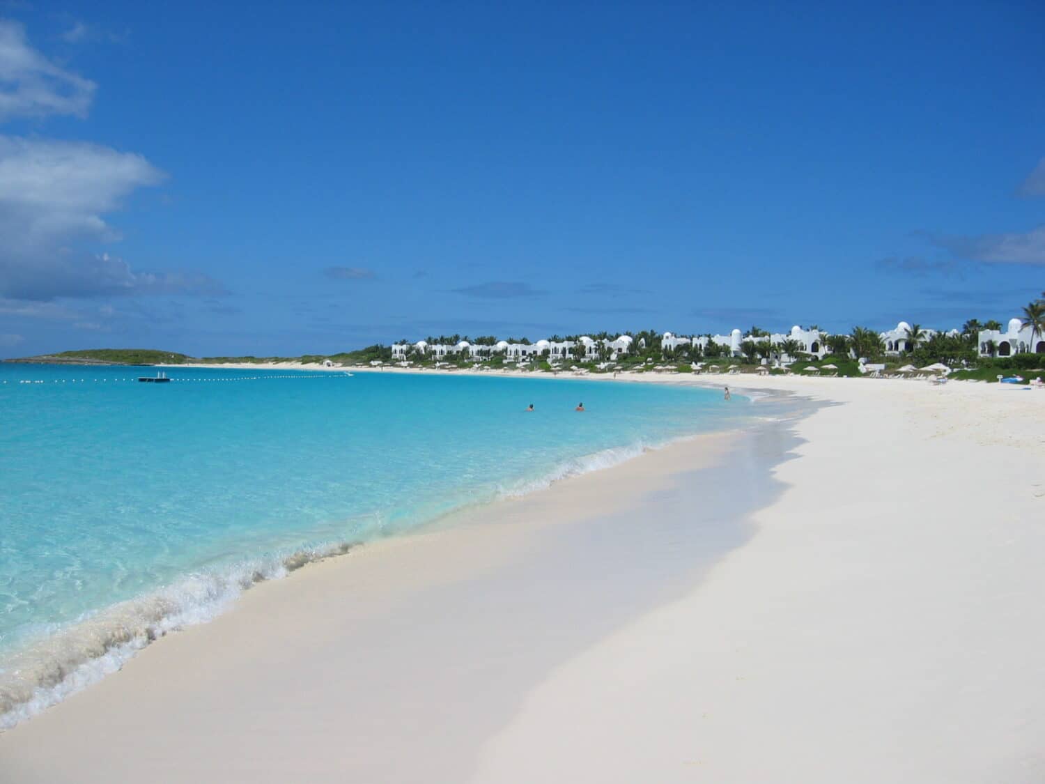 Praia com areia branca e mar azul cristalino, em um dia ensolarado, com o céu bem azul, além da vista de um resort, localizado na costa e rodeado de árvores verdes