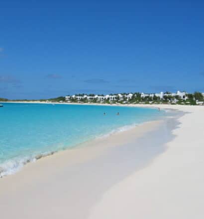Praia com areia branca e mar azul cristalino, em um dia ensolarado, com o céu bem azul, além da vista de um resort, localizado na costa e rodeado de árvores verdes