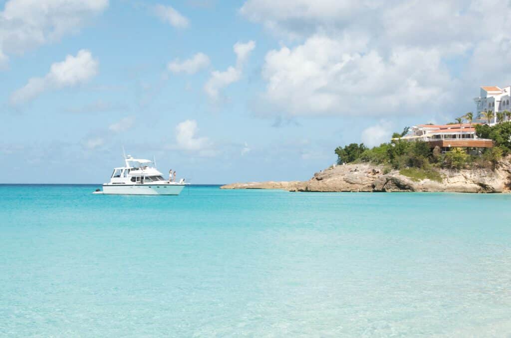 Mar cristalino, num tom de azul bem claro, durante um dia ensolarado na ilha de Anguilla, com um barco e pessoas olhando para o mar, além de uma parte costeira da ilha com um prédio branco em cima de uma rocha grande, rodeado de árvores verdes