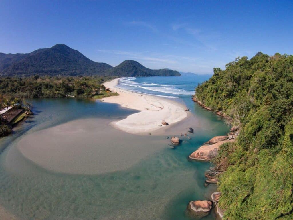 Praia da Itamambuca, um local de mar azul claro e esverdeado, a areia é bem clara e forma pequenas ilhas, do lado direito tem algumas pedras e montanhas