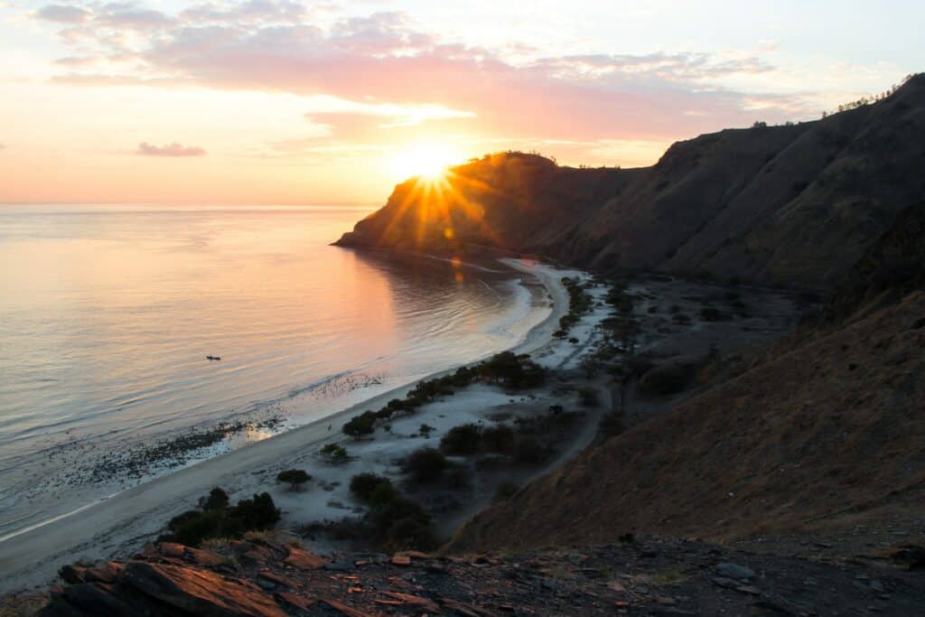 Praia em Timor Leste, águas azuis e uma grande faixa de terra com vegetação, o sol se pondo no horizonte e montanhas emoldurando a paisagem para representar o seguro viagem Timor Leste.