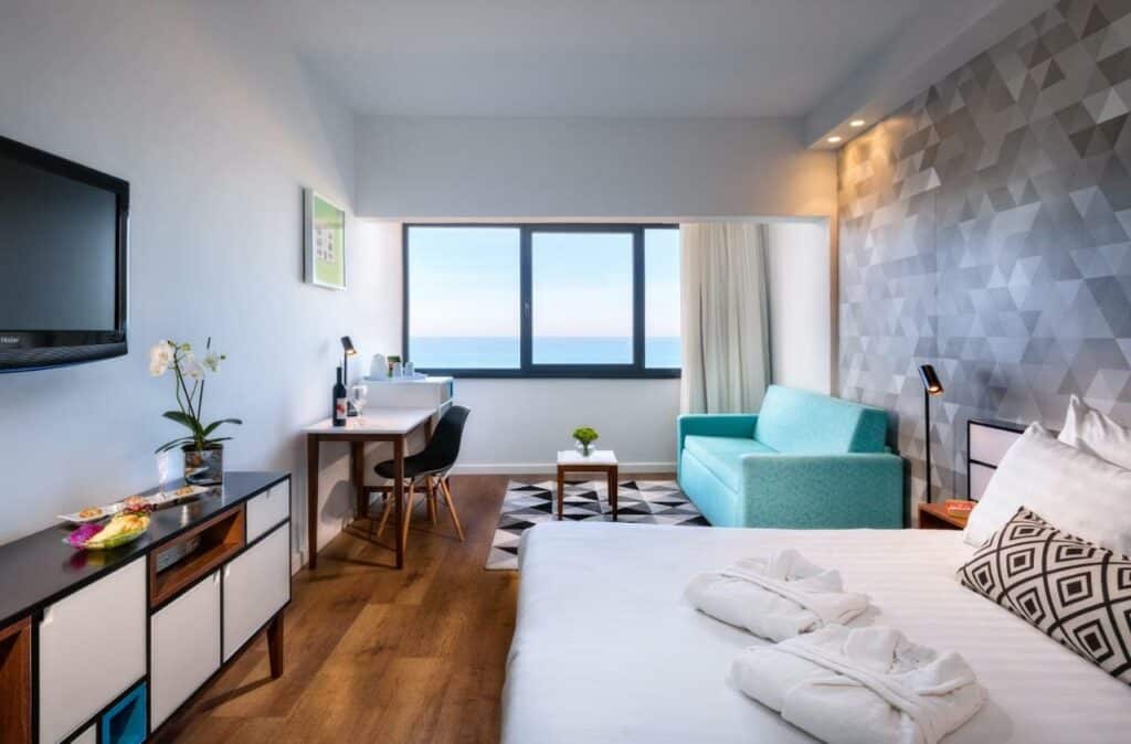 Quarto de hotel com cama de casal com lençóis brancos, toalhas brancas e travesseiros, um sofá azul de canto com televisão e aparador em frente ao sofá com janela com vista para o mar.