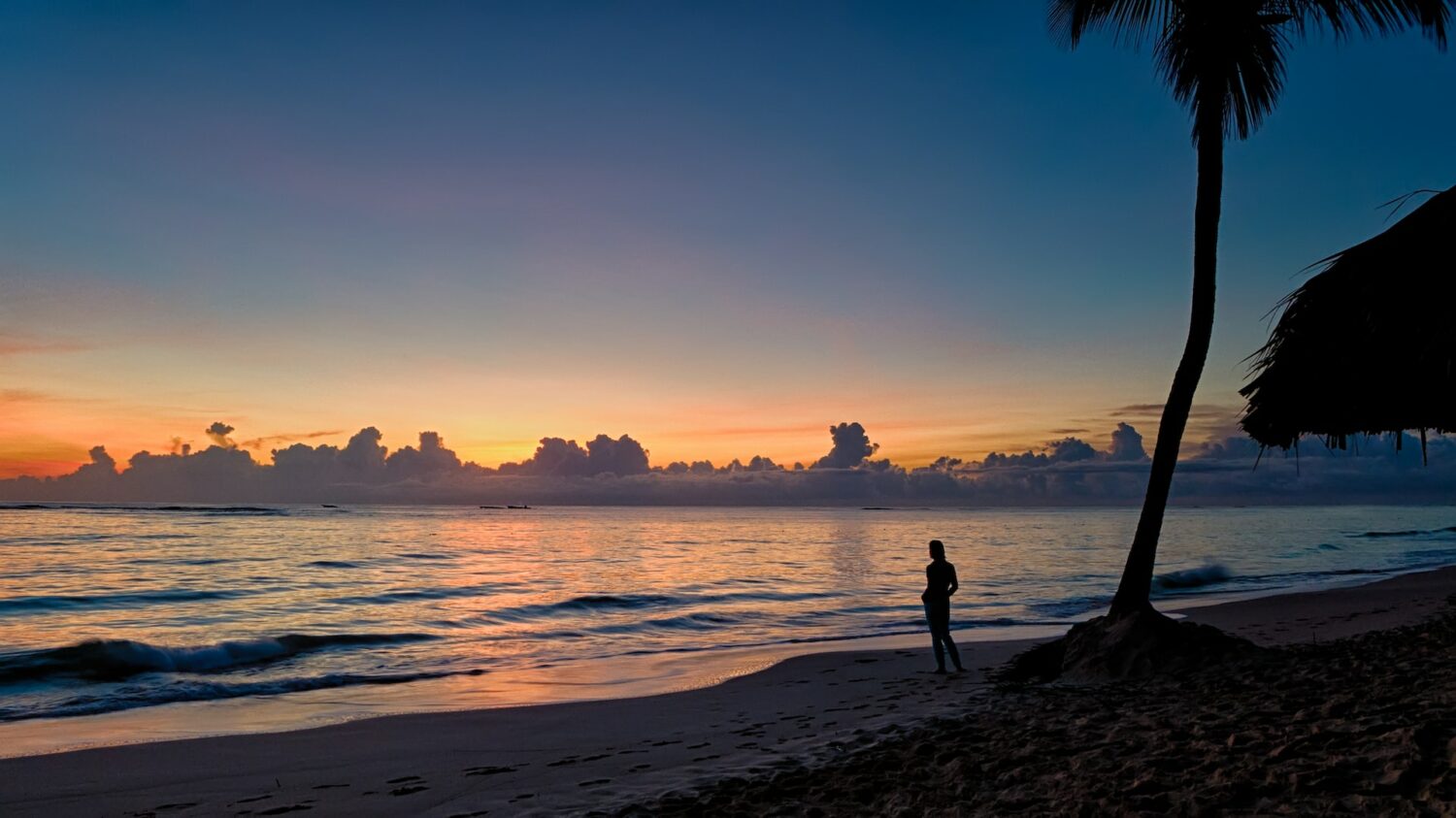silhueta de uma pessoa ao pôr do sol na praia de Punta Cana, para ilustrar o post de chip celular República Dominicana, há uma palmeira bem alta atrás da pessoa, e o mar é calmo com poucas ondas e reflete as cores alaranjadas do céu