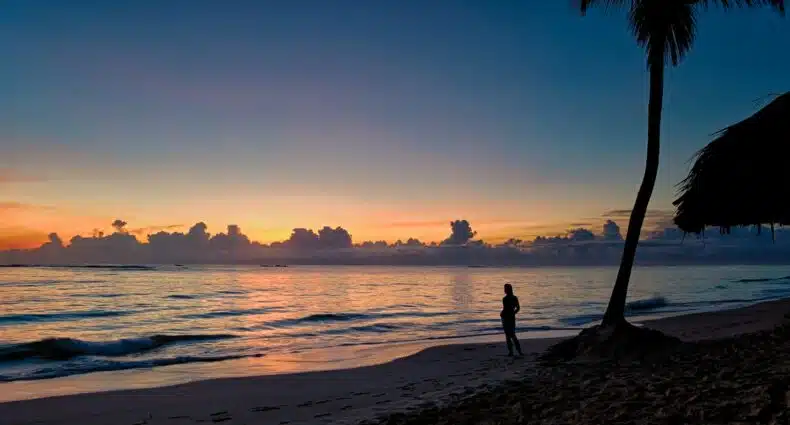 silhueta de uma pessoa ao pôr do sol na praia de Punta Cana, para ilustrar o post de chip celular República Dominicana, há uma palmeira bem alta atrás da pessoa, e o mar é calmo com poucas ondas e reflete as cores alaranjadas do céu