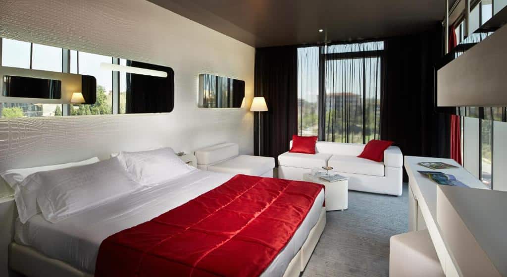 quarto grande do San Ranieri Hotel com cama king-size em detalhes vermelhos, dois sofás, sendo de um e dois lugares brancos, uma tv na frente e espelhos acima da cama, dos lados, luminárias e uma janela pegando toda a parede com veneziana e vista