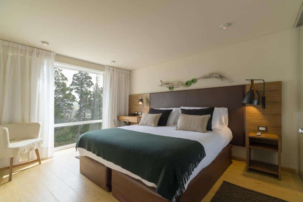 Quarto do Aguila Mora Suites & Spa com cama de casal, duas cômodas ao lado, cadeira branca do lado esquerdo e ao fundo janelas amplas com cortinas brancas.