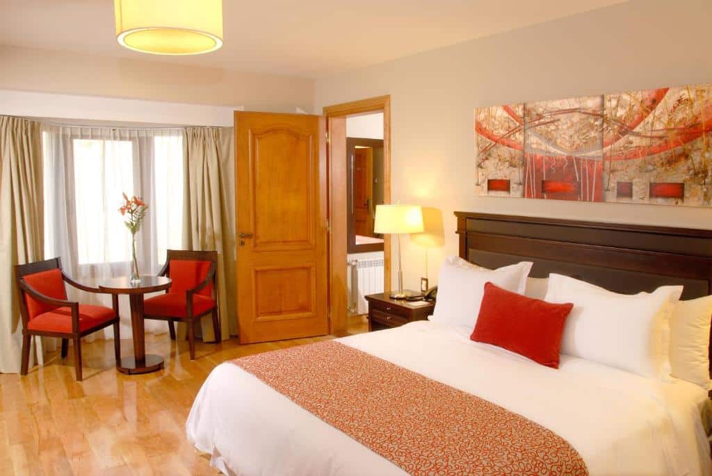 Quarto do Alma Del Lago Suites & Spa com cama de casal, do lado esquerdo cômoda de madeira com luminária e duas cadeiras estofadas de vermelho e mesa com vaso de planta perto da janela com cortinas beges.