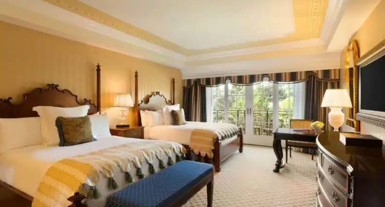 Quarto do Fairmont Grand Del Mar com duas camas de casal, sacada com vista para o jardim, cômoda de madeira em frente a cama com TV em cima. Representa hotéis em San Diego