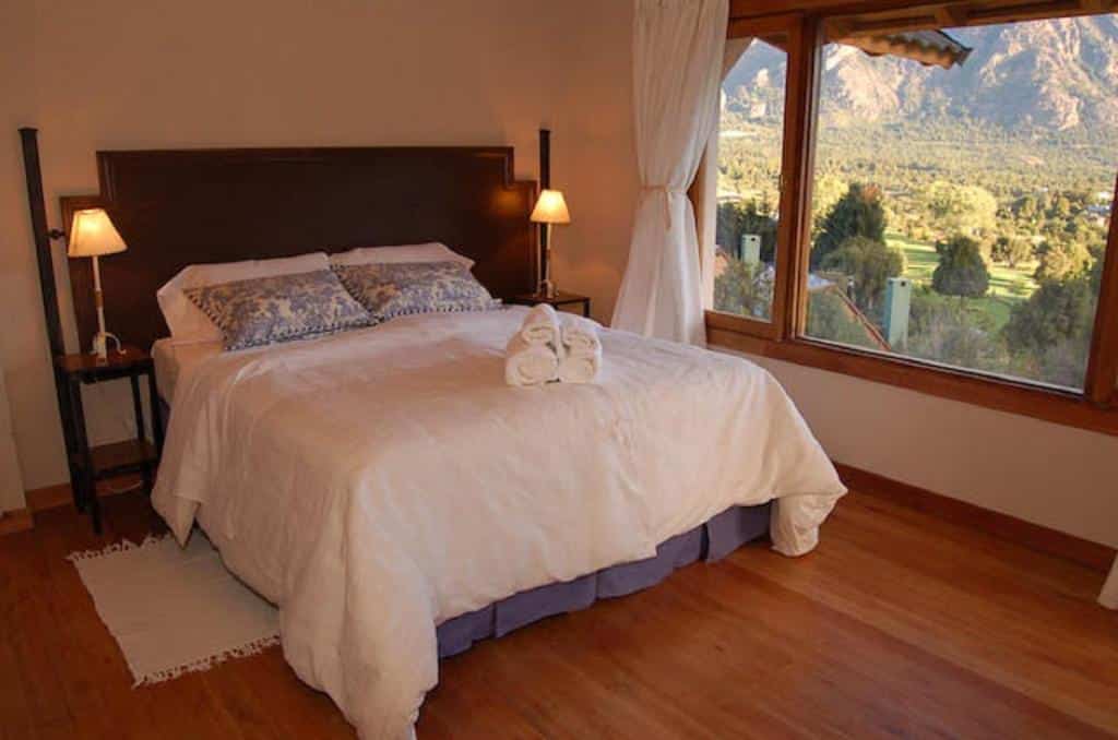 Quarto do Arelauquen Bungalows & Suites com uma cama de casal, duas cômodas do lado da cama com luminárias e do lado esquerdo tem uma janela ampla com cortinas brancas. Representa hotéis em Bariloche