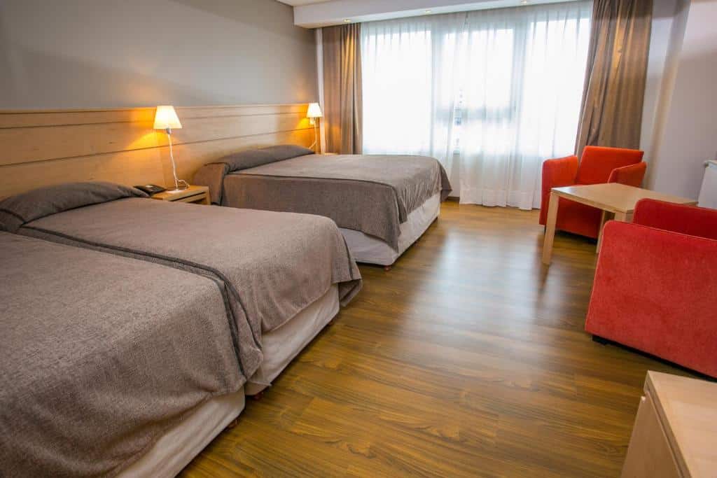 Quarto do Hotel Ayres Del Nahuel com duas camas de solteiro do lado direito, uma cômoda com luminária e do lado esquerdo tem uma cama de cala, janelas do lado esquerdo com cortinas brancas e em frente a cama de casal duas poltronas vermelhas e uma mesa de madeira no meio delas.