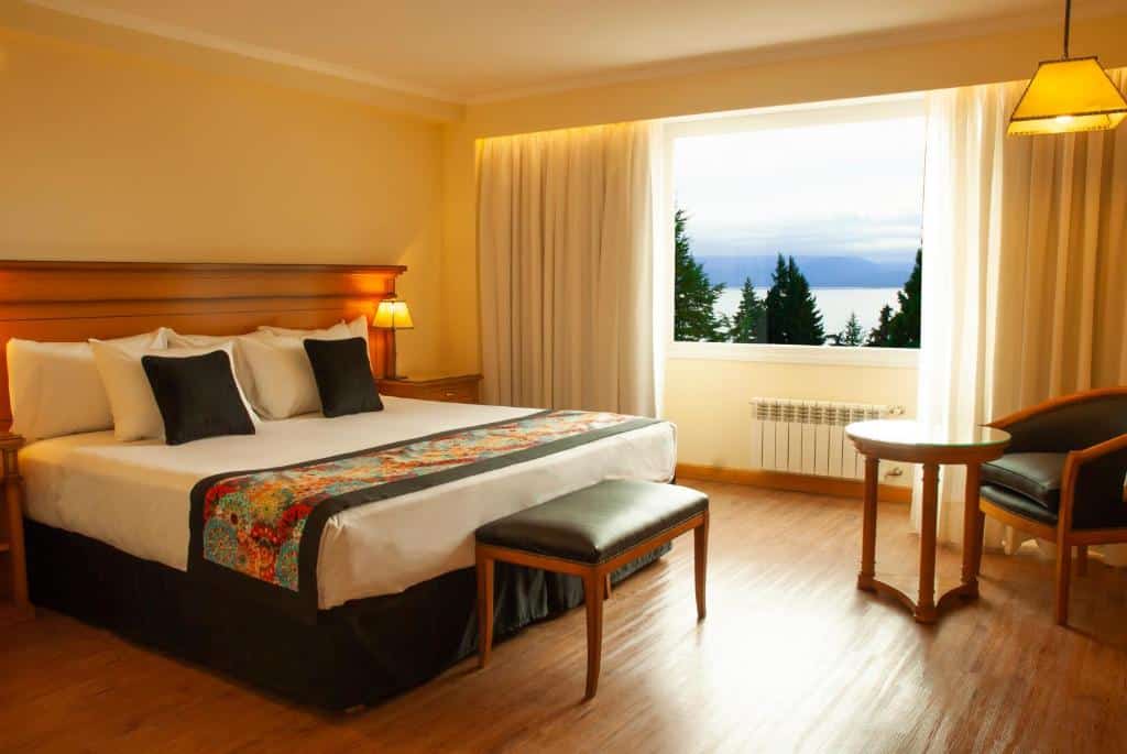 Quarto do Huinid Bustillo Hotel & Spa com cama de casal, uma cômoda do lado esquerdo com luminária, do lado esquerdo janela ampla com cortinas brancas, poltrona com mesa de madeira e na beira da cama banco estofado preto.