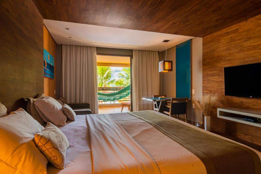 Quarto do Carmel Charme Resort com cama de casal ampla, TV em frente a cama, mesa com duas cadeiras do lado esquerdo, porta com cortinas brancas aberta com varanda com rede.