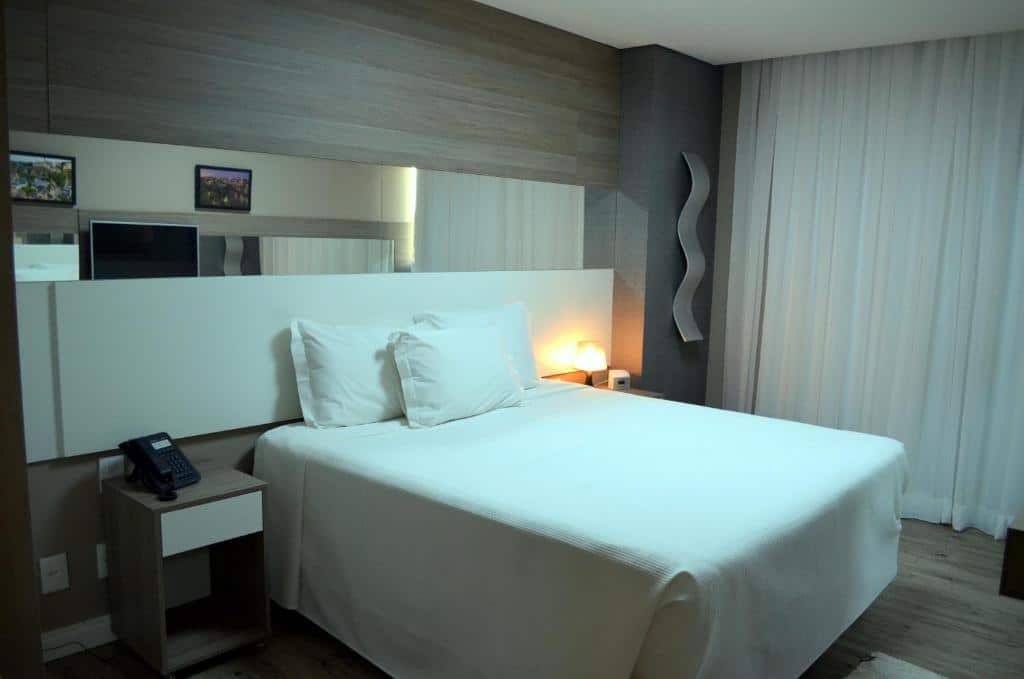 Quarto do Ville Celestine Condo Hotel e Eventos com cama de casal, duas cômodas ao lado, no lado esquerdo há uma iluminaria em cima e no lado direito um telefone.