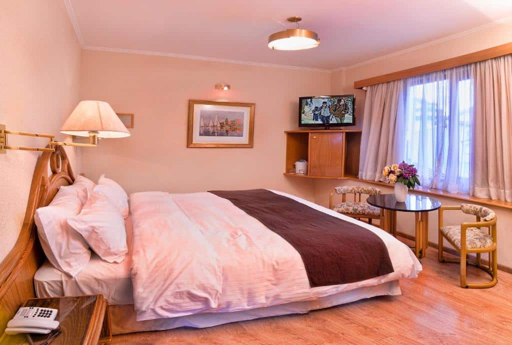 Quarto do Hotel Cristal com cama de casal, em frente a cama de casal mesa com duas cadeiras estofadas e ao fundo uma janela com cortinas brancas e do lado esquerdo no canto uma cômoda de madeira com TV em cima.