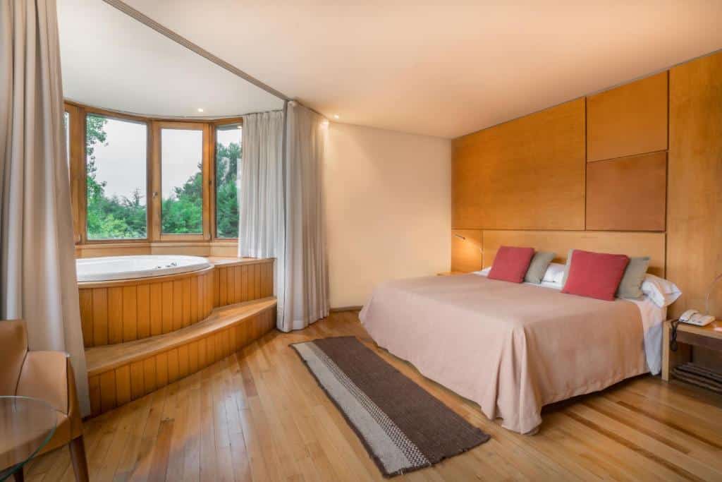 Quarto do Design Suites Bariloche com cama de casal, duas cômodas ao lado da cama e em frente da cama uma banheira de hidromassagem com janelas panorâmicas.