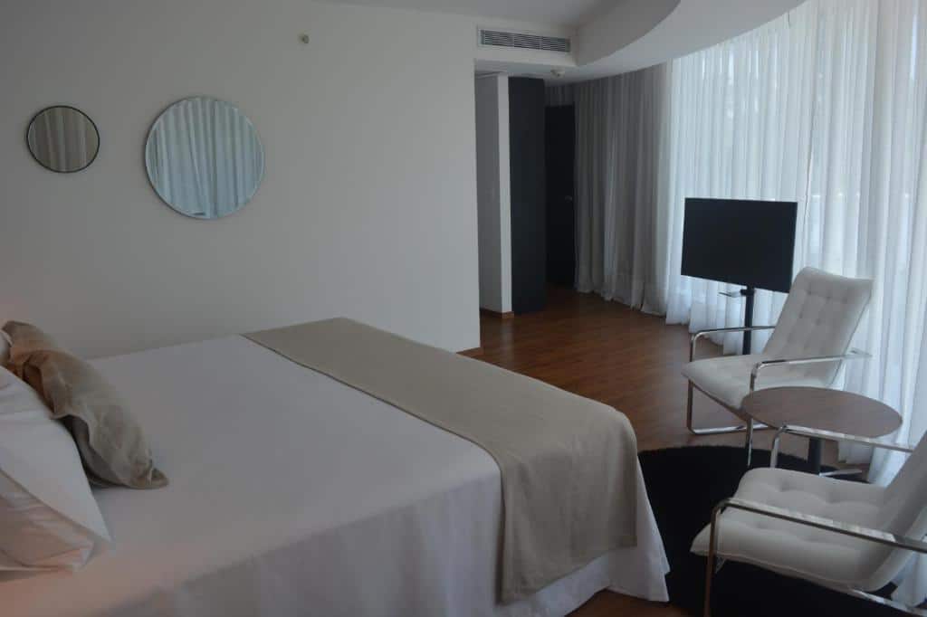 Quarto do Don Hotel  com cama de calsa, dois espelhos redondos na parede do lado direito, em frente a cama uma cadeira com uma TV.