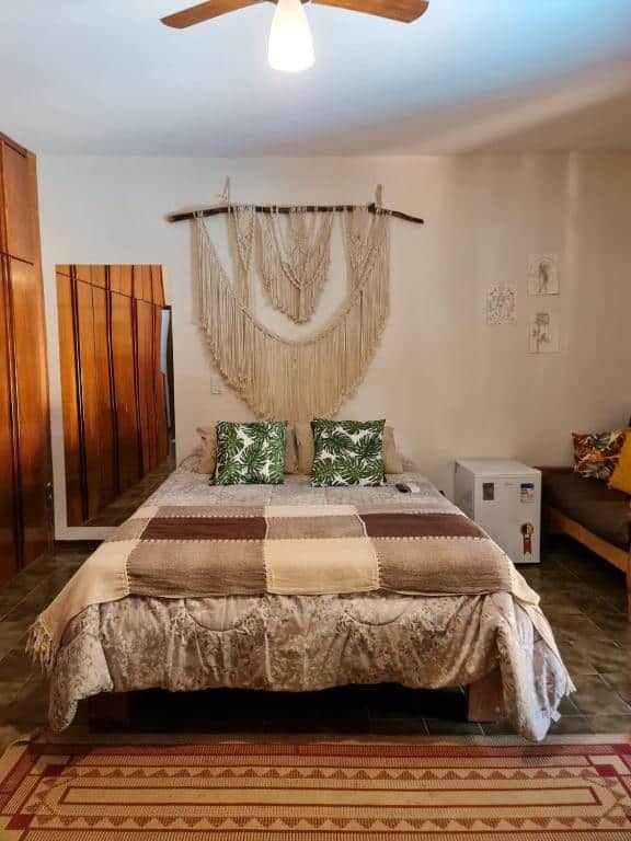 Quarto rustico do Ecocentro Brasil – Espaço Ecológico com cama de casal no centro do quarto, espelho grande e guarda roupa do lado direito espelho da cama e no lado esquerdo frigobar com um sofá.