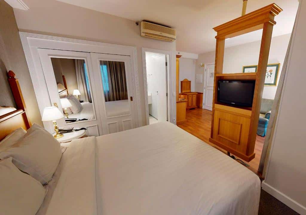 Quarto no Estanplaza Ibirapuera com uma cama de casal, um pequeno armário de duas portas com espelho, ar-condicionado, uma televisão e um outro cômodo com sofá, televisão, e o chão é de madeira