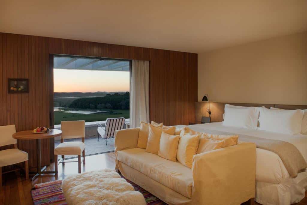 Quarto do Hotel Fasano Punta del Este com cama de casal, sofá na beira da cama, mesa com duas cadeiras perto da varada e  varanda com cadeiras.