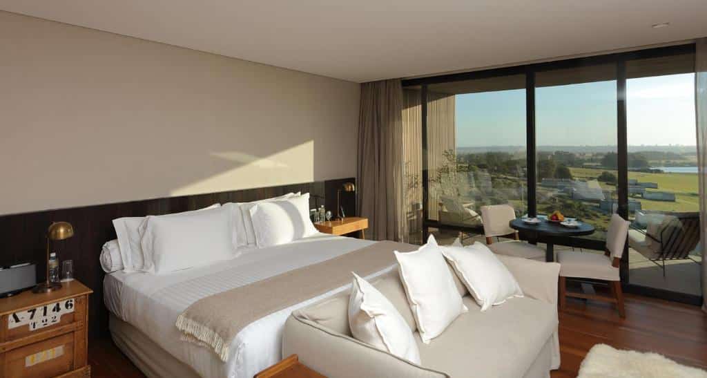 Quarto do Hotel Fasano Punta del Este com cama de casal, sofá em frente a cama, mesa com duas cadeira em frente a parede de vidro que dá acesso a sacada.
