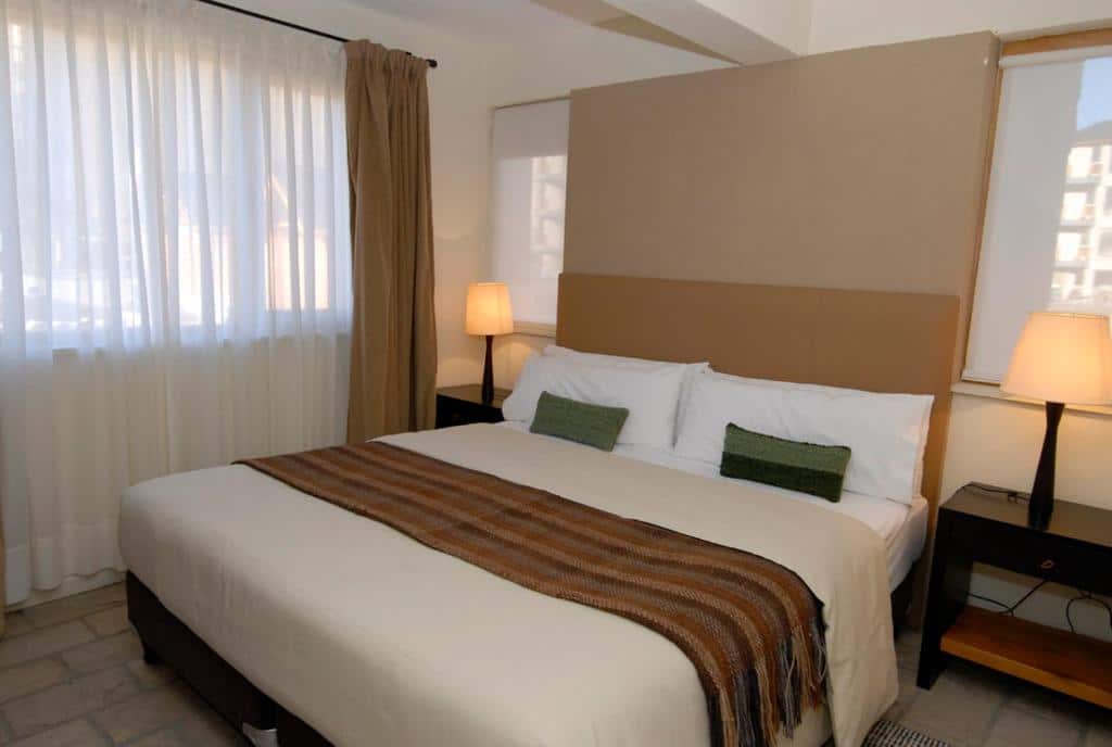Quarto do Galileo Boutique Hotel com cama de casal, duas cômodas ao lado de cama com luminárias e do lado esquerdo janelas com cortinas brancas.