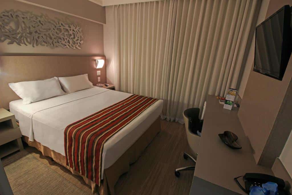Quarto do Hotel Beaga Convention Expominas by MHB com cama de casal, duas cômodas ao lado, janela com cortinas brancas do lado esquerdo, mesa de trabalho em frente a cama e na parede uma TV de tela plana.