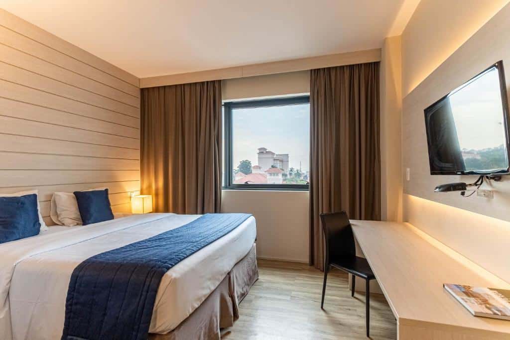Quarto no Hotel Contemporâneo - Royal Palm Hotels & Resorts com uma janela com cortinas, uma bancada ampla com uma cadeira, uma cama de casal, chão de madeira e uma televisão, para representar hotéis perto do Aeroporto de Viracopos em Campinas