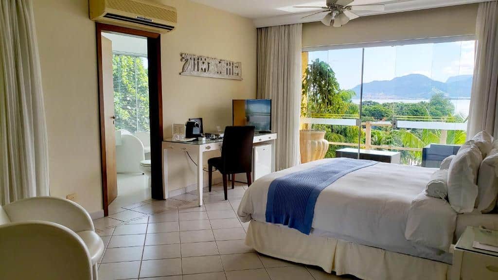 Quarto no Hotel Itapemar - Ilhabela com cama de casal, uma varanda com vista para o mar, uma poltrona, um frigobar e uma mesa de escritório com cadeira