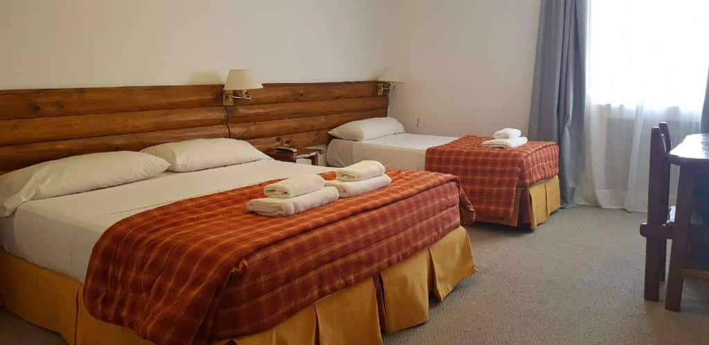 Quarto do Gran Hotel Panamericano com cama de casal, com toalhas em cima, uma cômoda ao meio separando de uma cama de casal do lado esquerdo em frente mesa de trabalho e do lado esquerdo janelas com cortinas brancas e azuis claras.