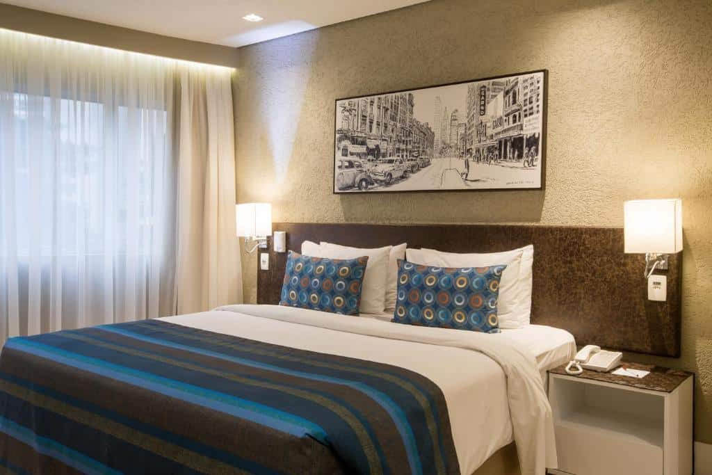 Quarto no Hotel Transamerica Berrini com uma cama de casal, uma janela com cortinas, cabeceira com iluminação indireta e um quadro sob a cama, para representar hotéis perto do Aeroporto de Congonhas