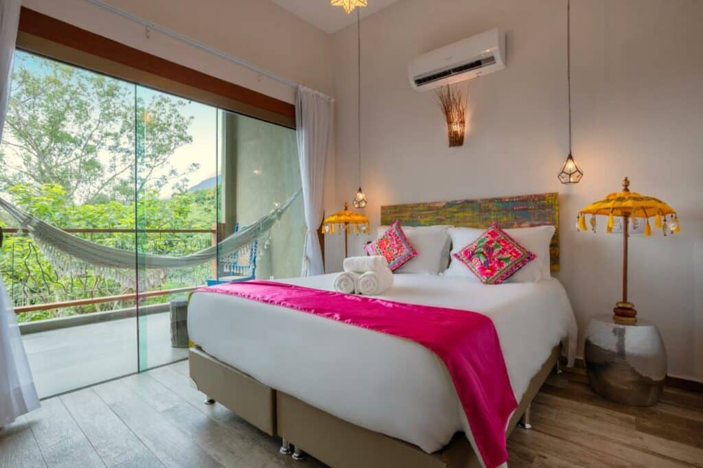 Quarto no Hotel Vila Kebaya com uma cama de casal, chão de madeira, uma varanda com rede, itens de decoração rústicos pelo quarto, para representar resorts em Ilhabela