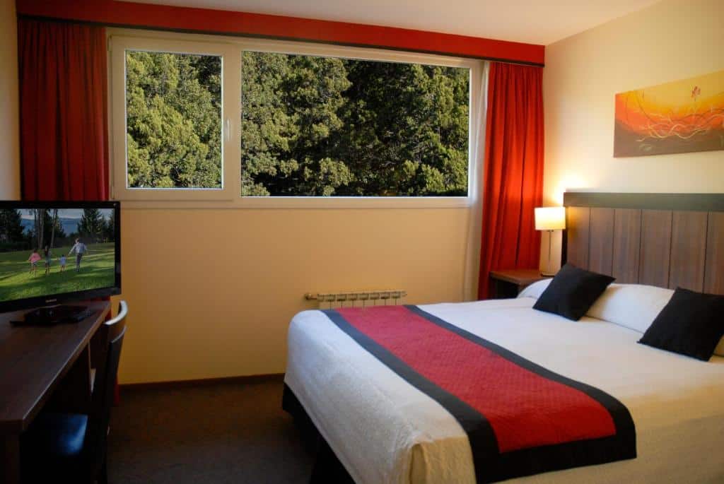 Quarto do Huinid Pioneros Hotel com cama de casal, uma cômoda do lado esquerdo perto da janela com cortinas laranjas. Em frente a cama, tem uma cômoda de madeira com cadeira e uma TV em cima.