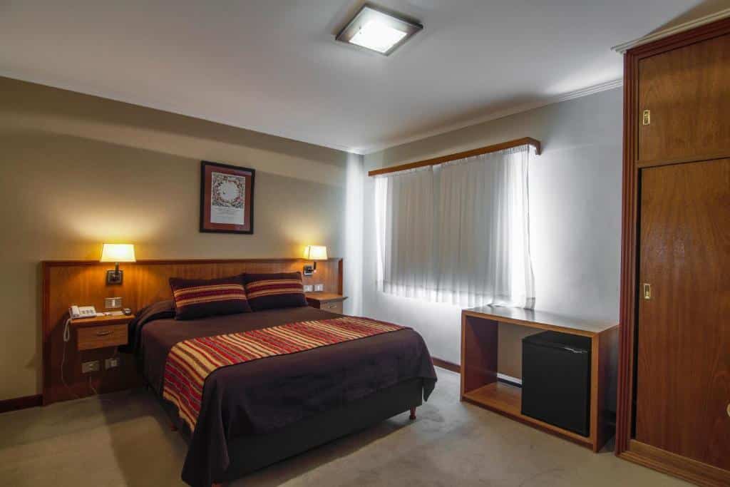 Quarto do Hotel Milan com cama de casal, duas cômodas ao lado com luminária, mesa com frigobar do lado esquerdo e ao fundo uma janela com cortinas brancas.