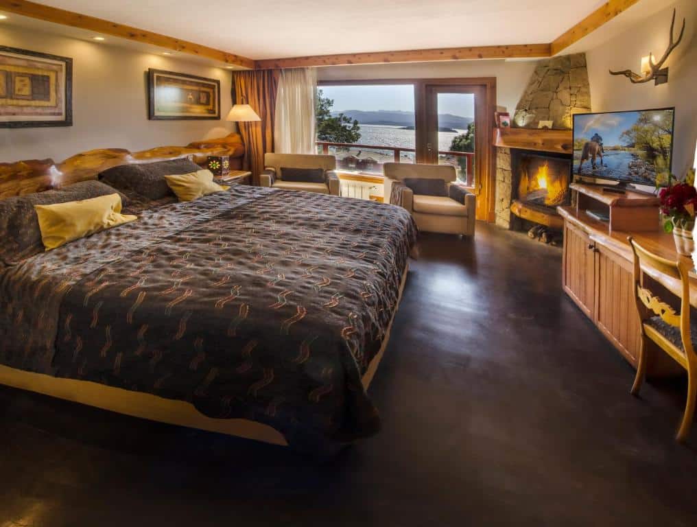 Quarto do Nido del Cóndor Hotel & Spa com cama de casal, duas poltronas do lado esquerdo em frente a portas de vidro que dá acesso a varanda. Em frente a cama TV em cima de uma cômoda de madeira.