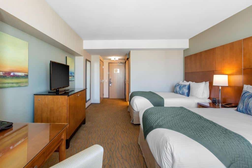 Quarto do Omni San Diego Hotel com duas camas de solteiro e uma cômoda com luminária no meio delas. TV em frente as camas em cima de uma cômoda de madeira e mesa de trabalho com cadeira no lado esquerdo.