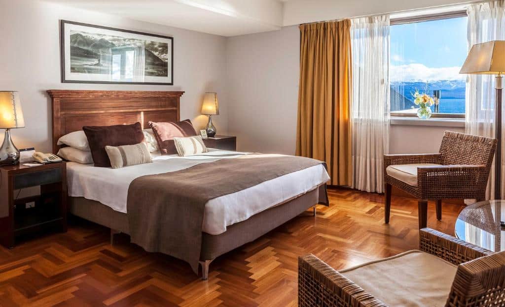 Quarto do Hotel Panamericano Bariloche, com cama de casal, duas cômodas ao lado com luminárias em cima, duas cadeiras de frente a cama e uma janela esquerdo com cortinas brancas e amarelas, com vista do mar