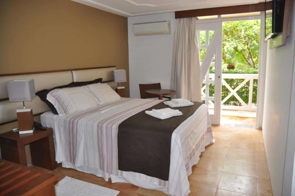 Quarto do Hotel Porto Belo com cama de casal, duas cômodas ao lado, mesa com cadeira do lado esquerdo perto da varanda.