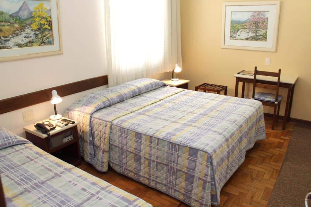 Quarto do Hotel São Bento com mesa com cadeira no lado esquerdo, cama de casal com duas cômodas com iluminarias ao lado e uma cama de solteiro do lado direito.