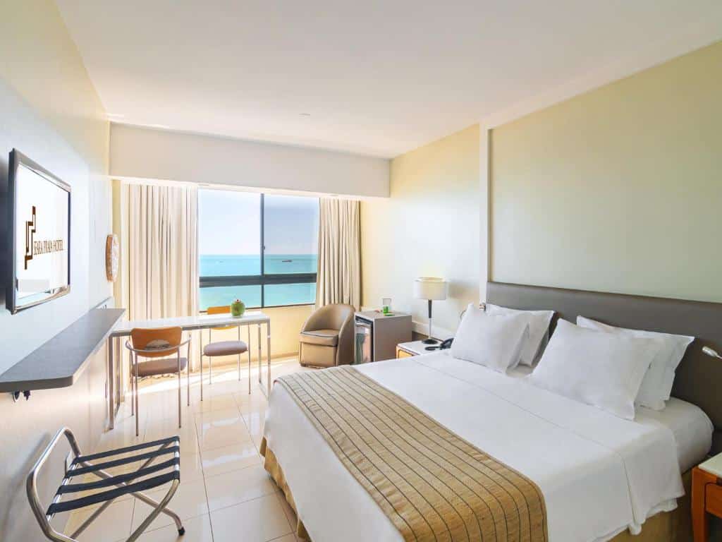 Quarto do Seara Praia Hotel com cama de casal, TV em frente cama, mesa do lado esquerdo perto da janela e poltrona ao lado do frigobar.