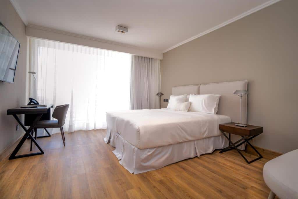 Quarto do Hotel Solerios  com cama de casal, duas cômodas com luminárias ao lado da cama, TV em frente a cama com mesa de trabalho com cadeira abaixo da TV.