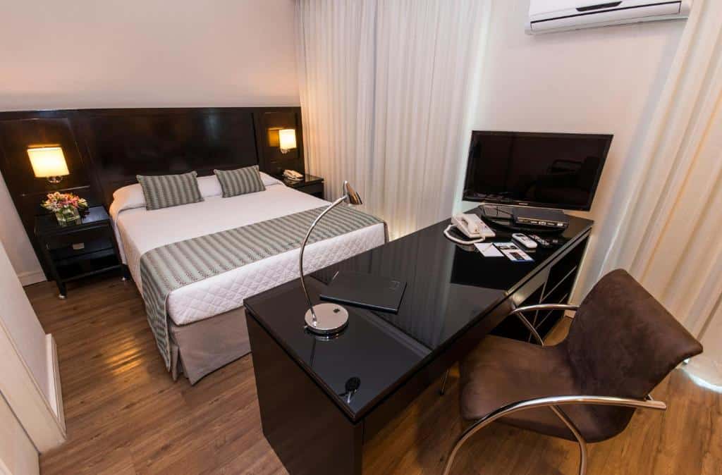 Quarto do Transamerica Executive Perdizes com uma cama de casal, chão de madeira, janelas com cortinas, ar-condicionado e uma mesa de escritório com uma cadeira. Esse é um dos hotéis perto do Allianz Parque.